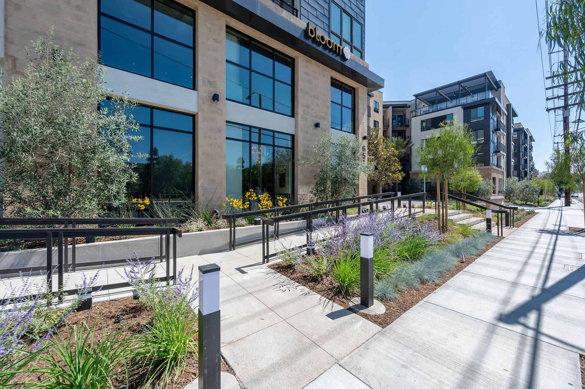 An exterior view of Bloom Apartments South Coast, with a sidewalk and lush greenery in the surroundings.