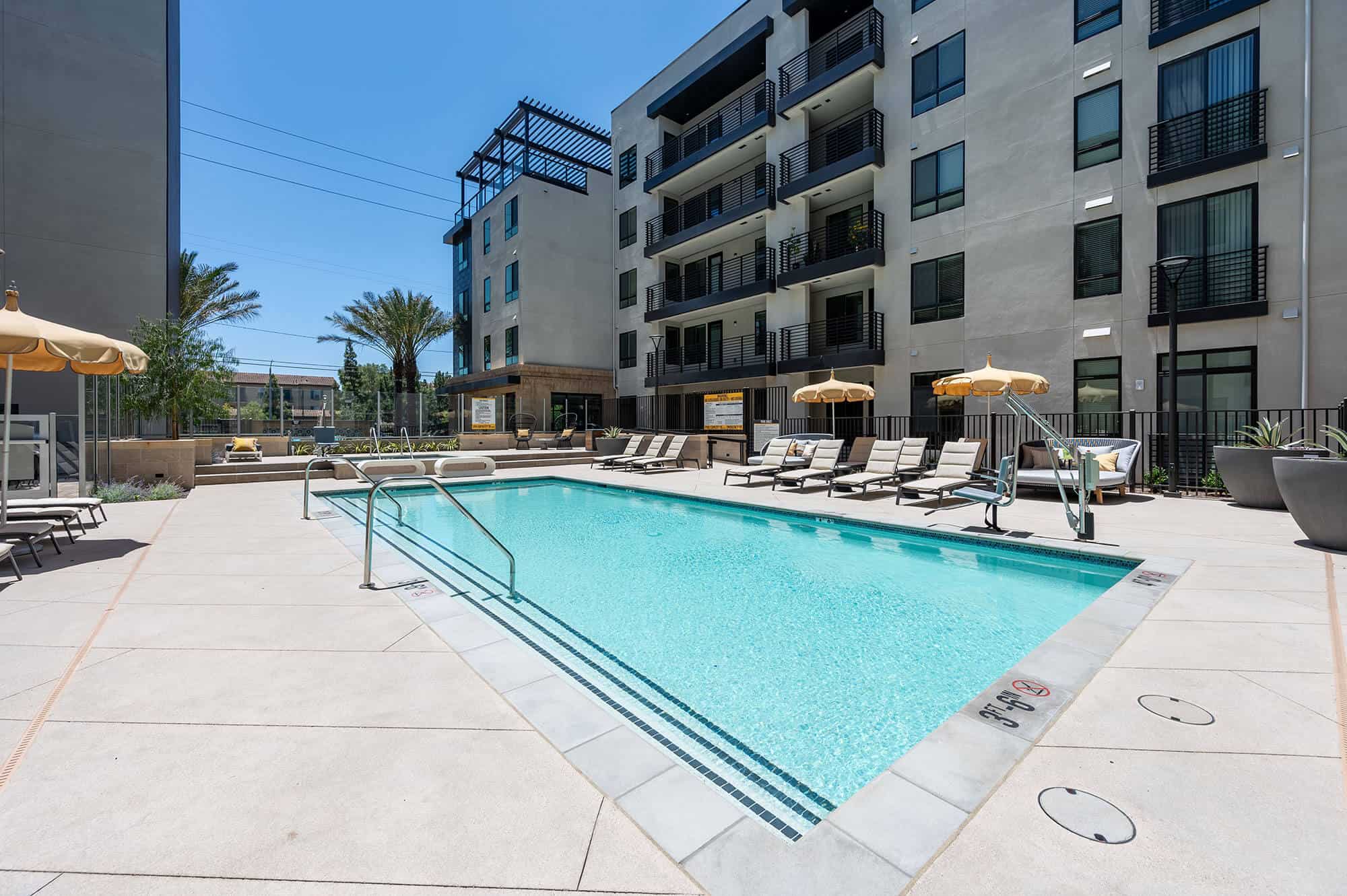 A view of the outdoor swimming pool area with lounge chairs, outdoor seating, and umbrellas for shade, with Bloom Apartments serving as the backdrop