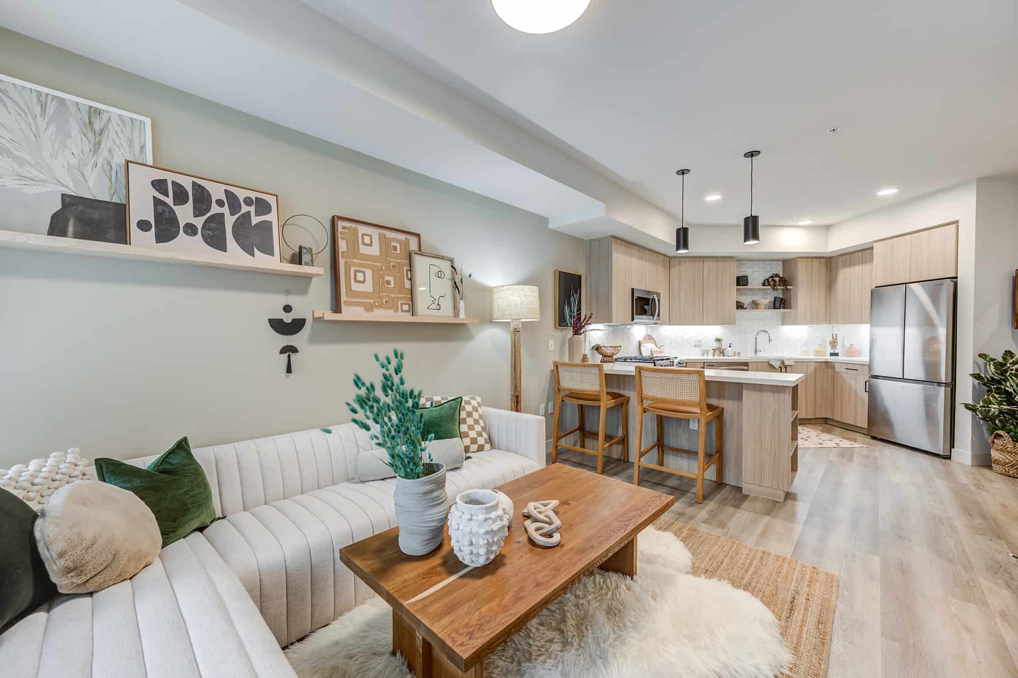 A living room featuring a white sofa and a wooden center table, alongside a kitchen area equipped with two high chairs and a refrigerator