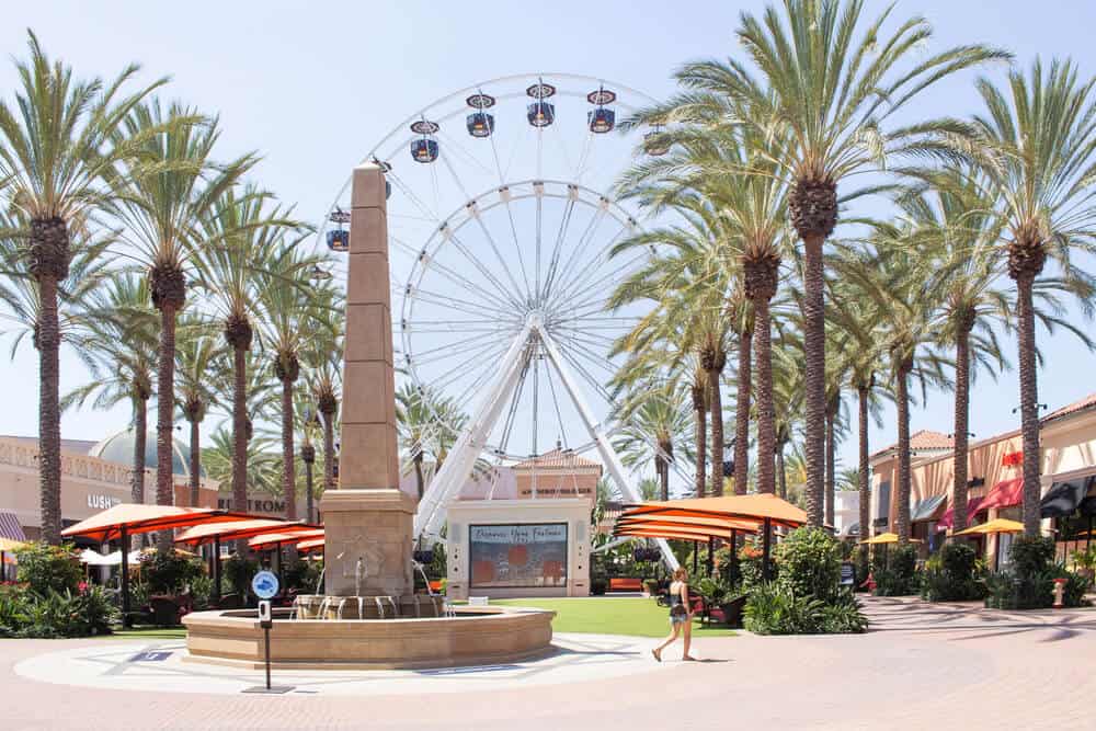 A white Ferris wheel rises behind a stone obelisk fountain in an outdoor shopping plaza lined with tall palm trees, with orange shade umbrellas over seating areas and a pedestrian walking across the sunlit pavement.