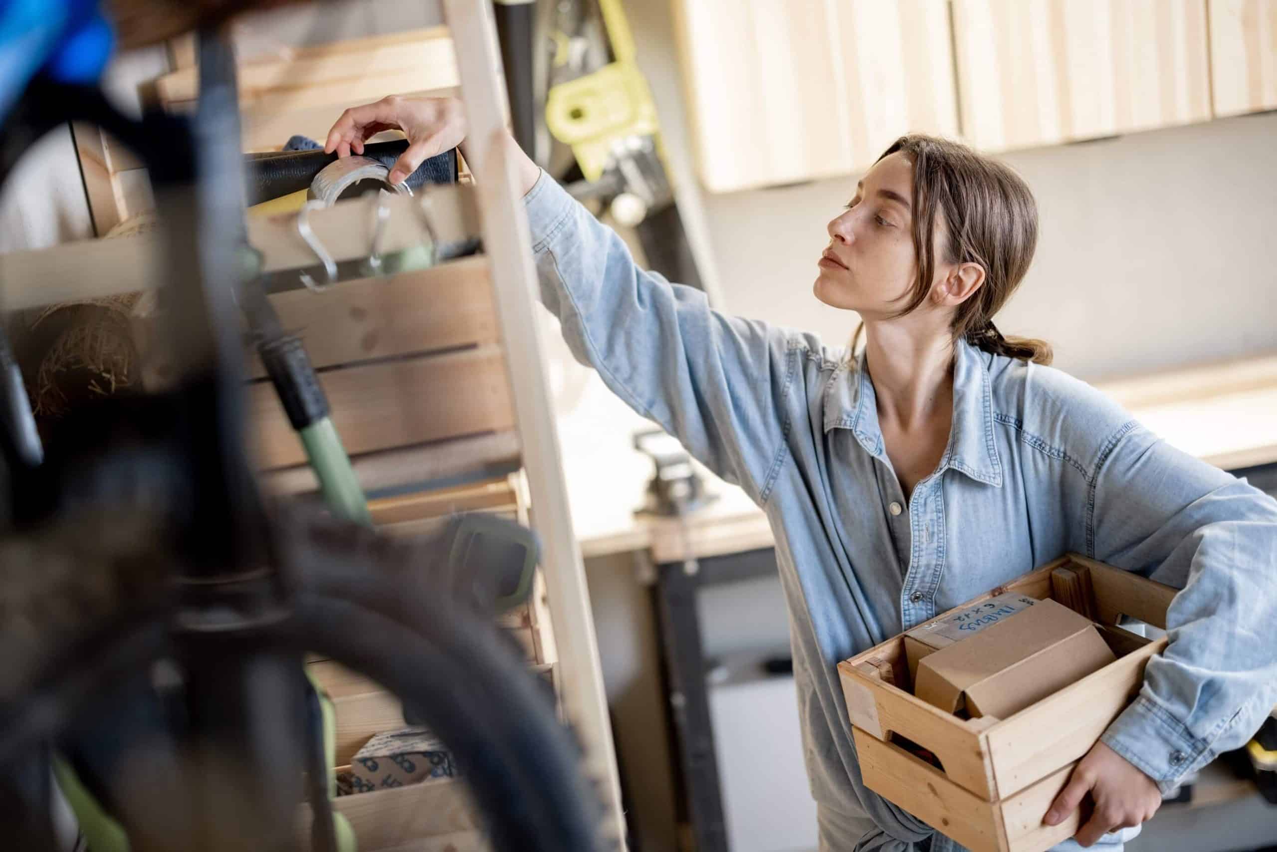 Woman in a denim shirt holding a wooden crate of small boxes while reaching up to check items on a garage storage shelf, surrounded by tools and a bicycle.